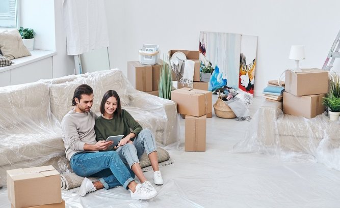 Happy young female and her husband sitting on rolled carpet by couch and scrolling through online furniture shop